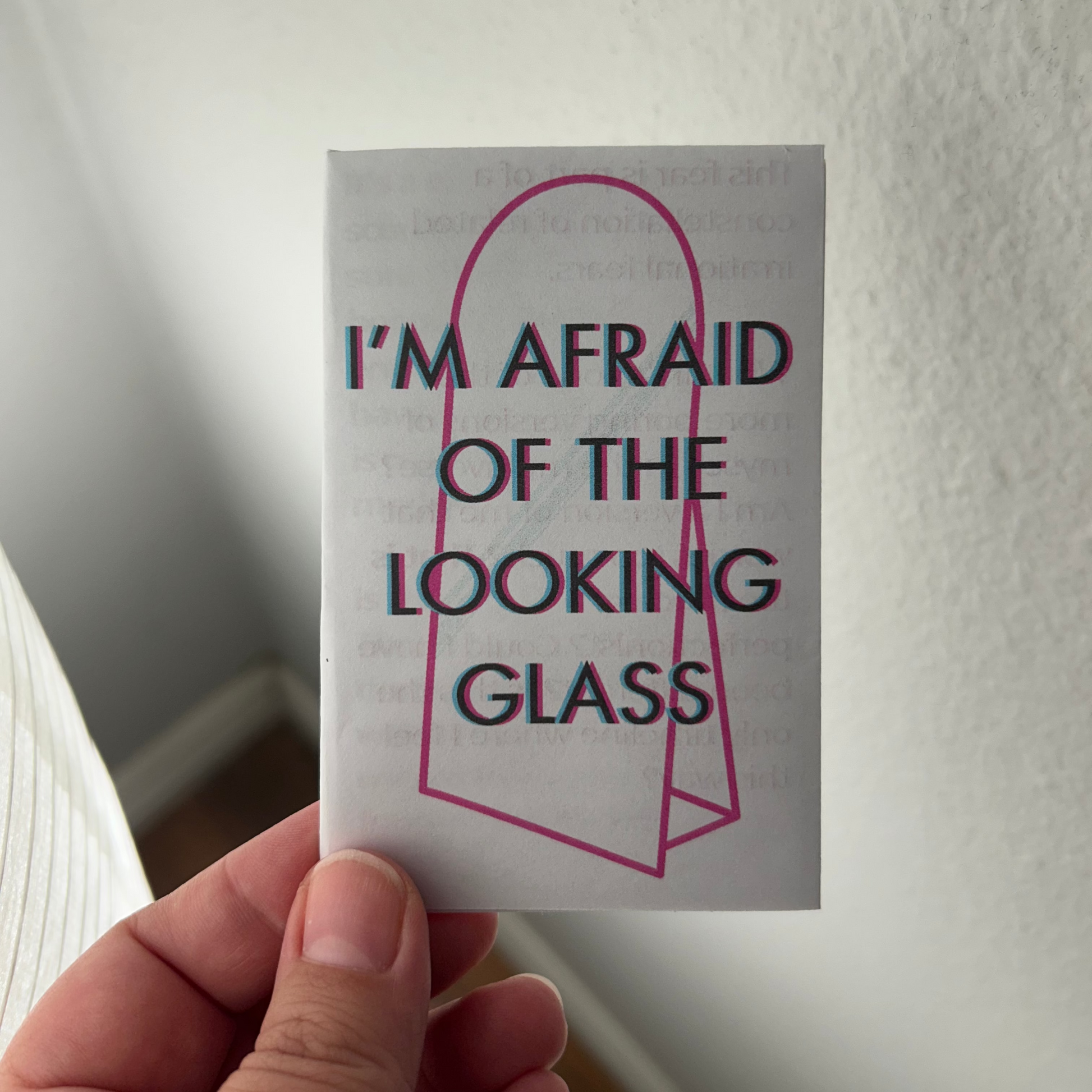 Hand holding a zinewith text 'I'm afraid of the looking glass' against a neutral background